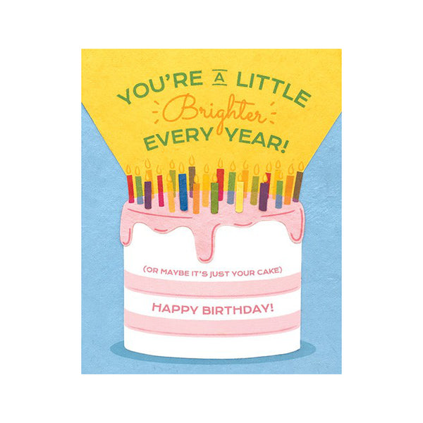 Fair Trade handmade card depicting a large white and pink cake with many candles on blue background. Yellow beam from candles reads "You're a little brighter ever year!" Message on cake reads "(or maybe it's just your cake) Happy birthday!