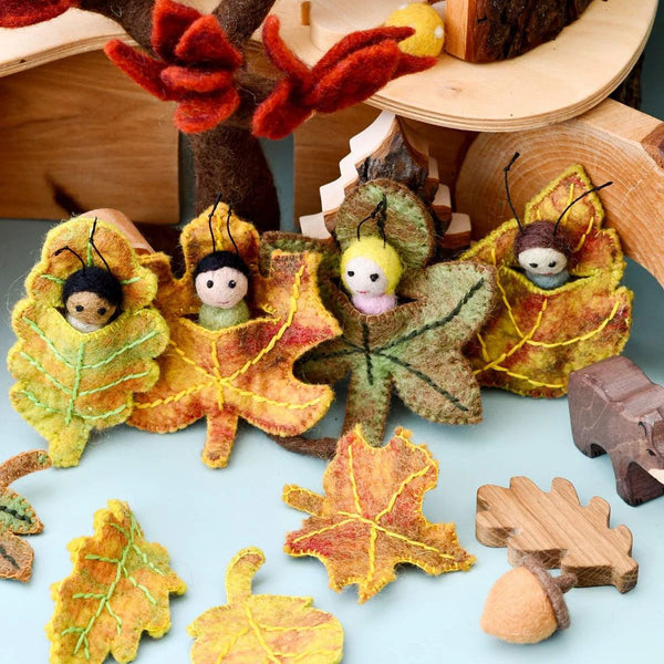 Felted leaf characters with small dolls inside, surrounded by autumn leaves and wooden elements on a light blue background.